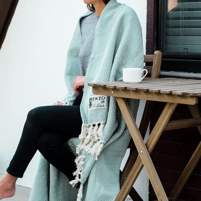 Woman sitting at table with white coffee cup on top wearing black pants and a green blanket 